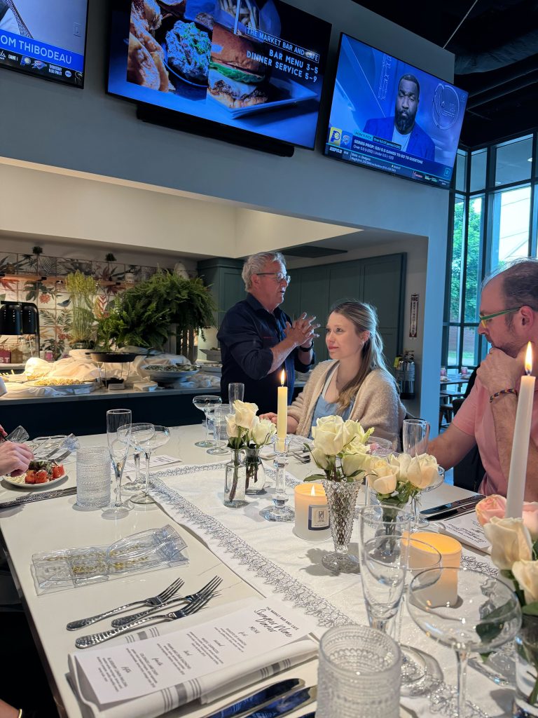 A wine dinner event at The Market Bar and Café. Pieter Ferreira is addressing seated guests at a white-decorated table adorned with white roses, candles, and elegant place settings.