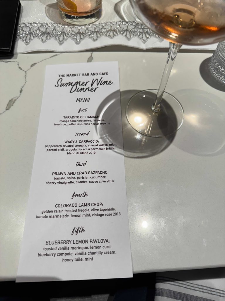The printed menu for "The Market Bar and Café Summer Wine Dinner" rests on the table under a wine glass. It lists five courses with descriptions and wine pairings, including taradito of hamachi, wagyu carpaccio, prawn and crab gazpacho, Colorado lamb chop, and blueberry lemon pavlova.