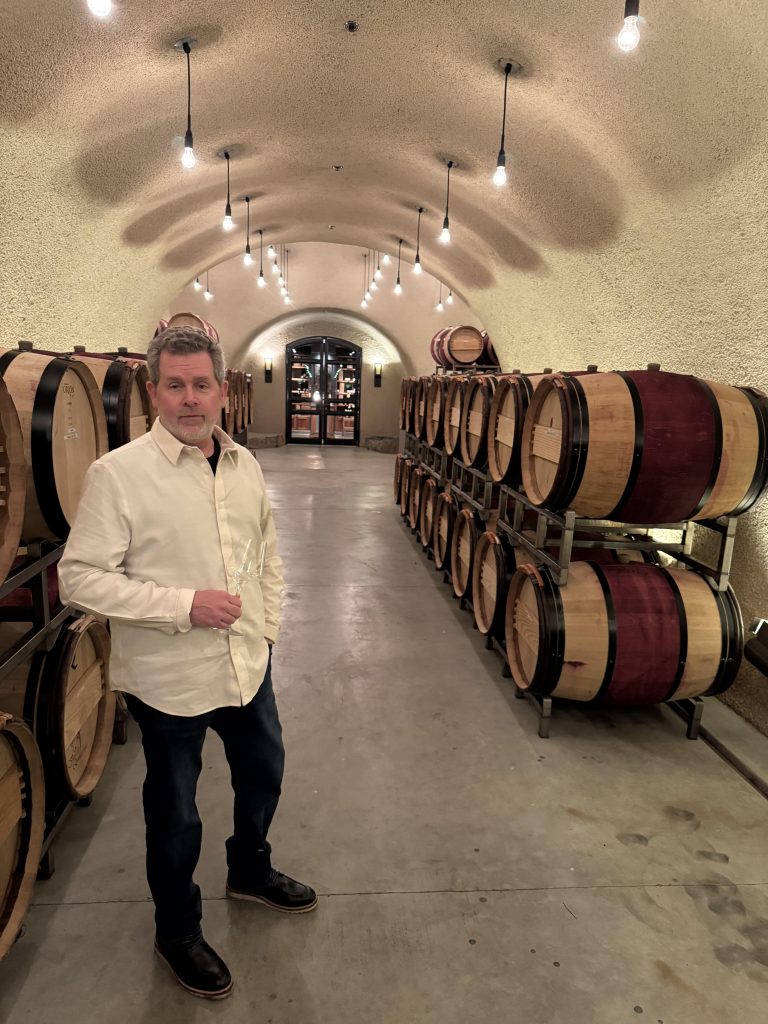 Chris posing in OVID Winery's curved concrete barrel cave with stacked oak barrels and a circular chandelier glowing in the background