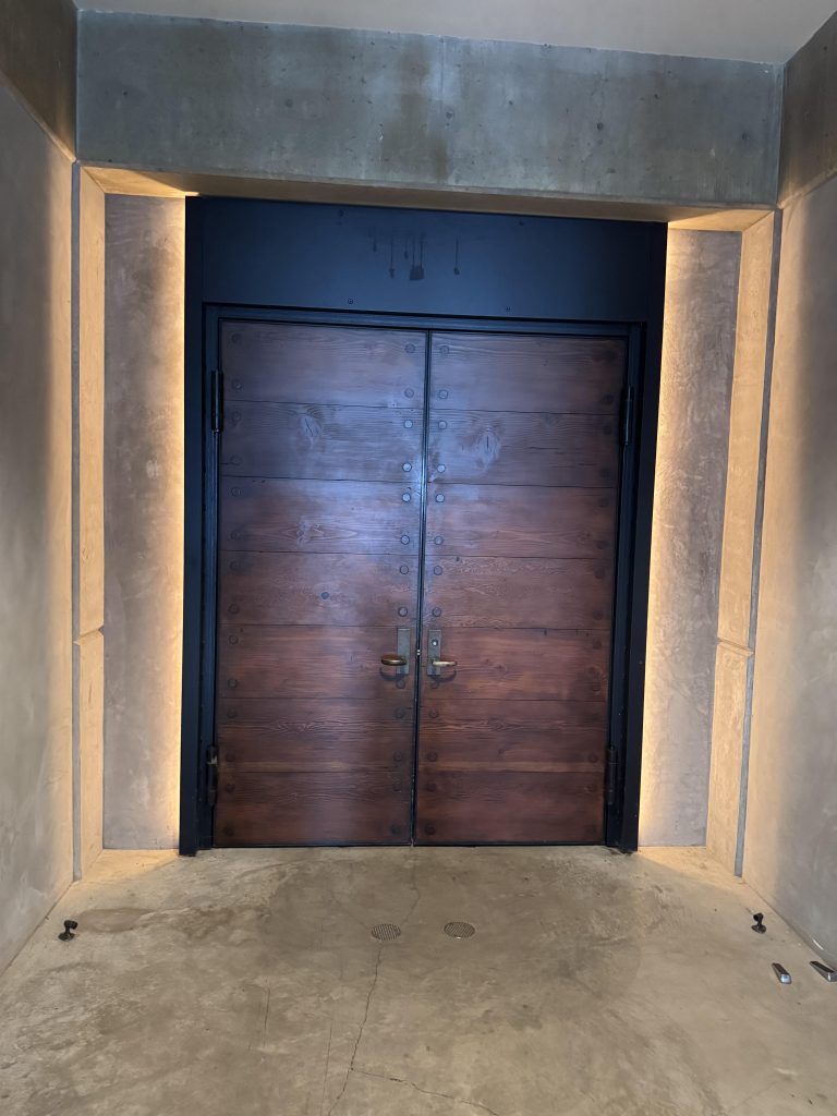 Dramatic upward-angle view of the heavy reclaimed wood entrance door at OVID Winery, framed in raw concrete with warm ambient underlighting