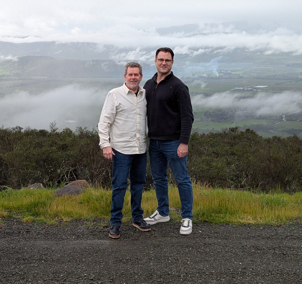 Chris and Nick posing on the hillside overlook at OVID Winery with a dramatic misty Napa Valley panorama behind them