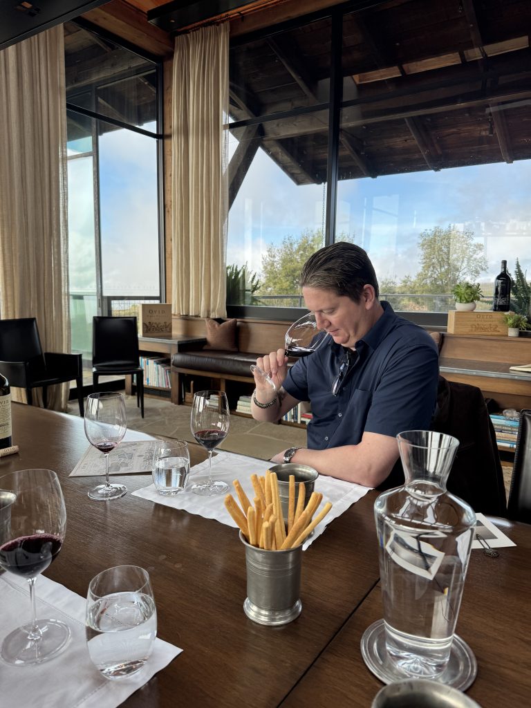 Matt nosing a glass of OVID at the tasting table, floor-to-ceiling windows framing the Napa hillside behind him