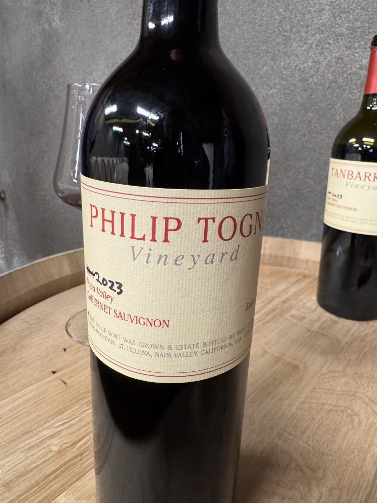Close-up of a Philip Togni Vineyard 2023 Cabernet Sauvignon bottle label resting on an oak barrel in the cellar