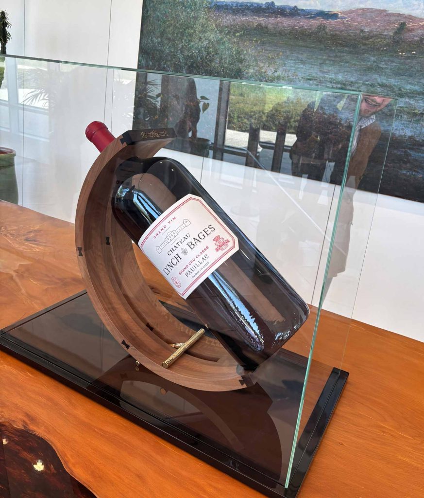 Large format bottle of Château Lynch-Bages resting on a decorative wooden cradle with a vineyard landscape painting visible behind it