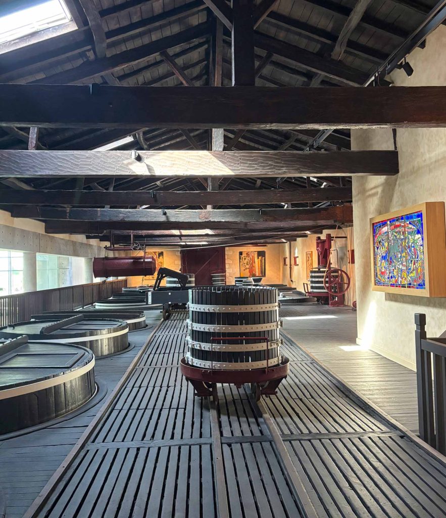 The historic original winery at Lynch-Bages featuring dark timber rafters, an old basket press, and a stained glass ceiling panel